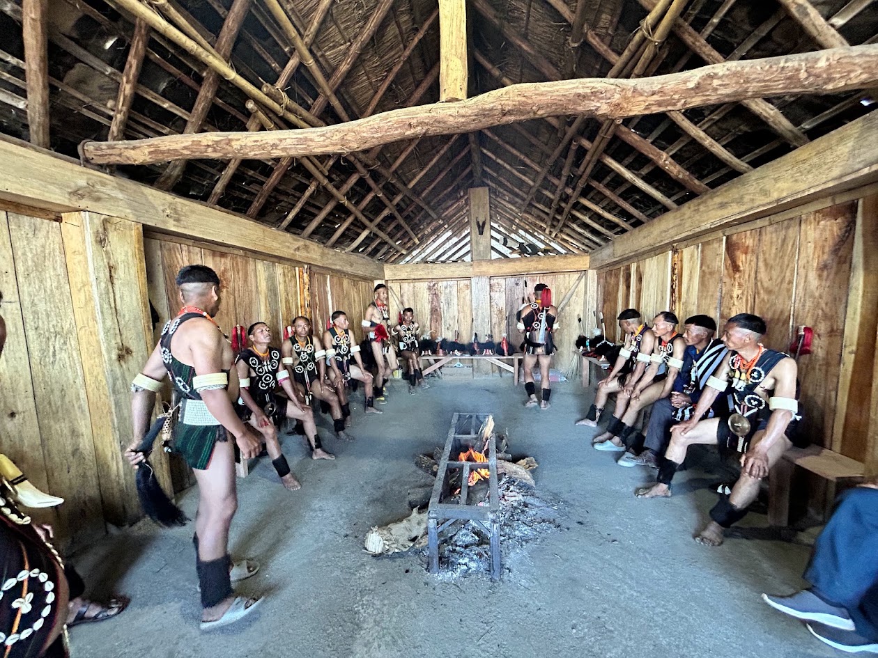 People in traditional clothing gathered around a hearth inside a wooden morung
